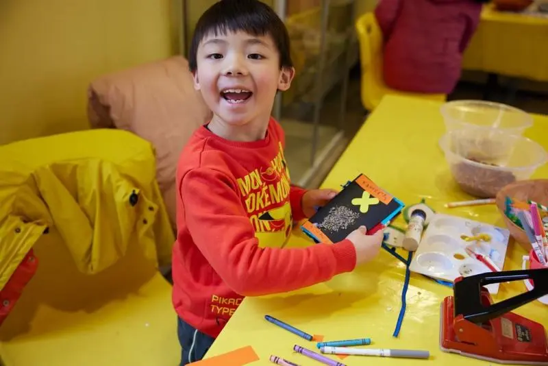 A very happy little boy shows the crafts he’s making on a yellow clothed table.
