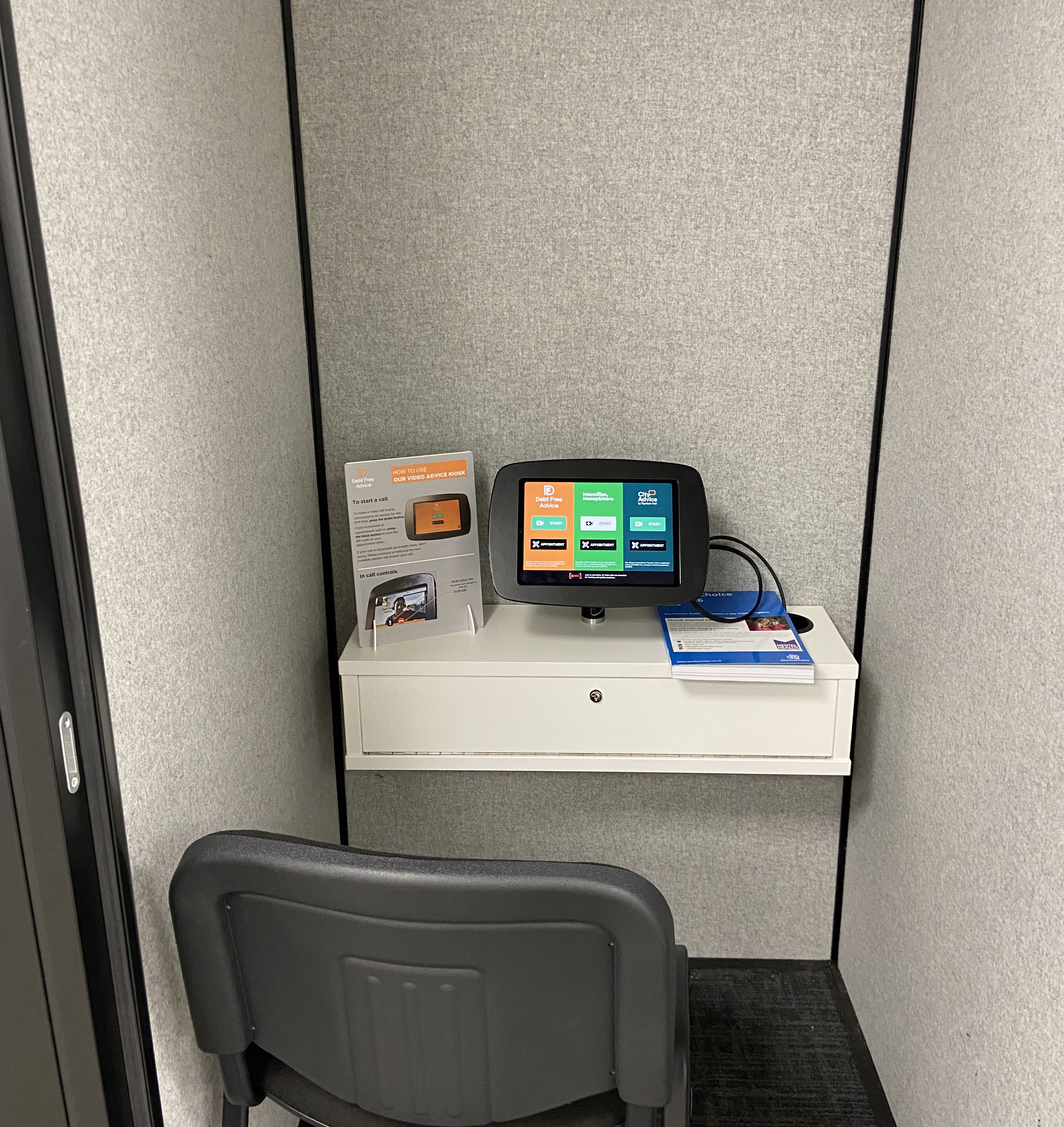 Inside the City Advice Kiosk booth with a small touchpad and leaflets dispenser on a ledge behind a chair.